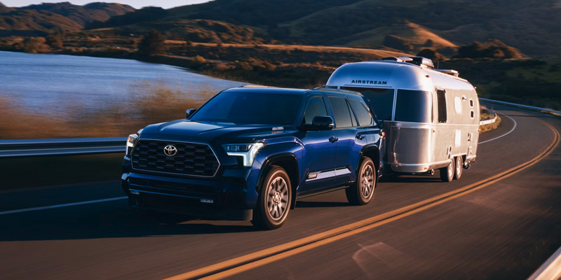 2025 Toyota Sequoia towing a silver Airstream trailer on a winding road near a body of water and mountains.