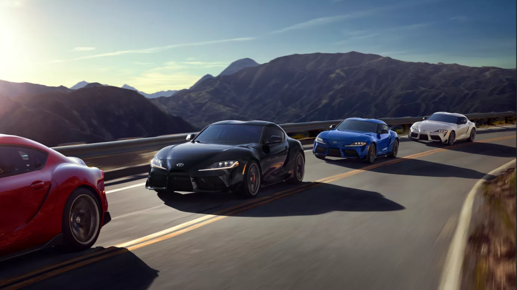 Five 2025 GR Supras in various colors driving on a winding mountain road at sunset