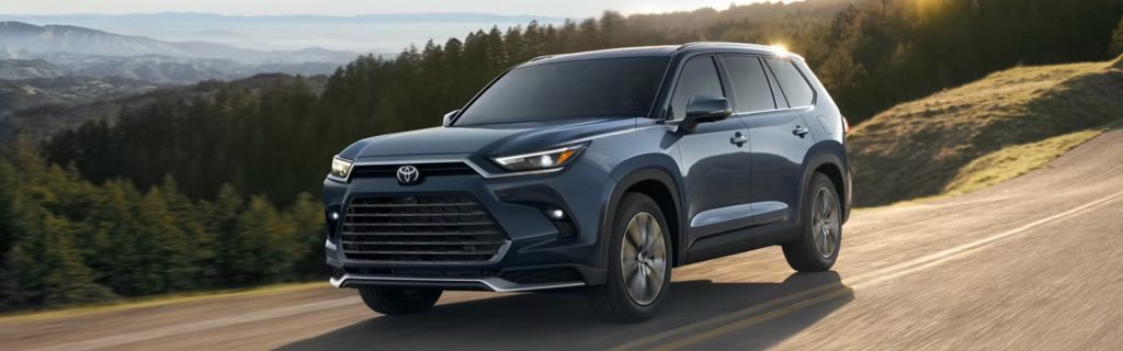 A dark blue Toyota SUV drives along a winding mountain road at sunset, with forested hills and layered mountains in the background.